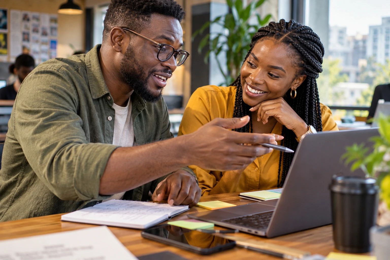 Two colleagues at a café desk with a laptop, one explaining with a pen while the other listens, smiling.